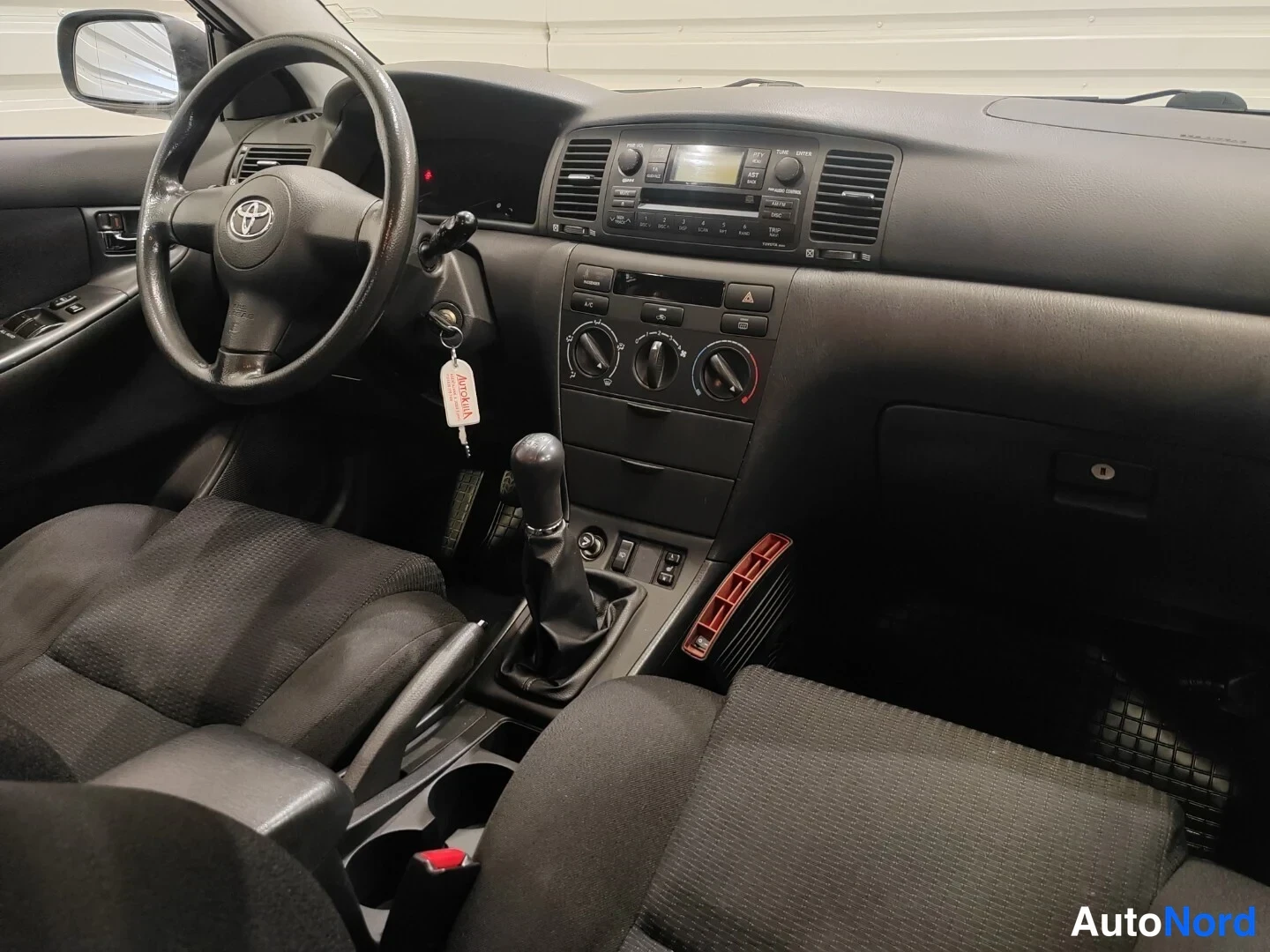 Muu: Toyota Corollan sisätilat kuljettajan puolelta: tumma verhoilu, ohjauspyörä, manuaalivaihteisto ja keskikonsoli.