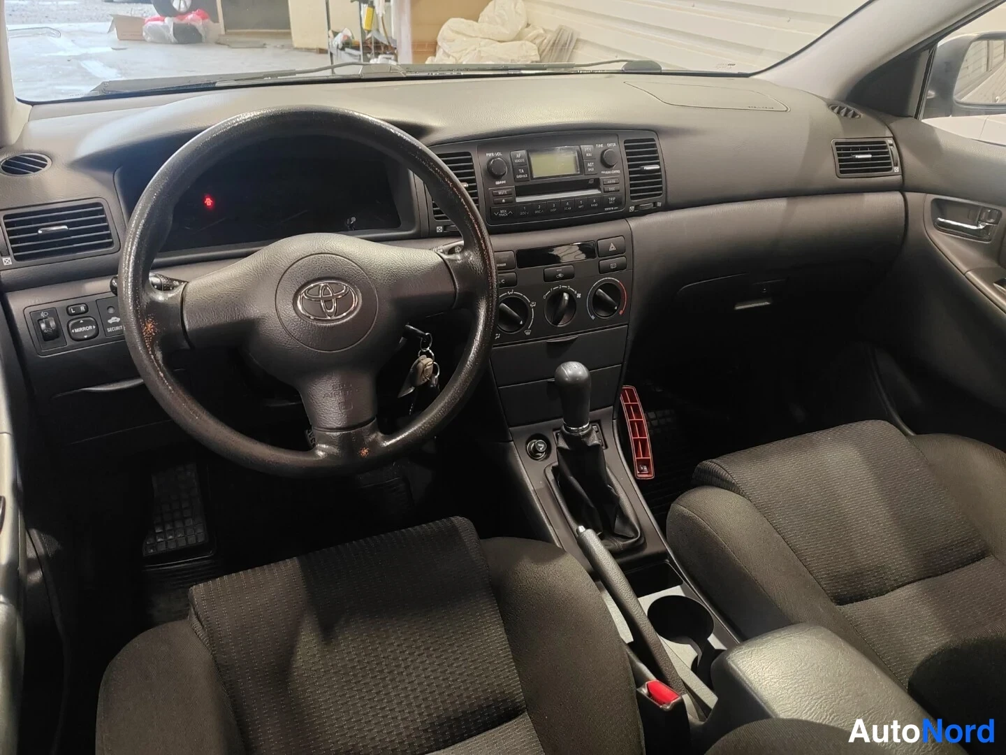 Etuviisto: Toyota Corollan ohjaamo etupuolelta: tumma tekstiiliverhoillut istuimet, Toyota-merkkinen ohjauspyörä, radio- ja ilmastointilaitteet keski-istuimessa, viisivaih