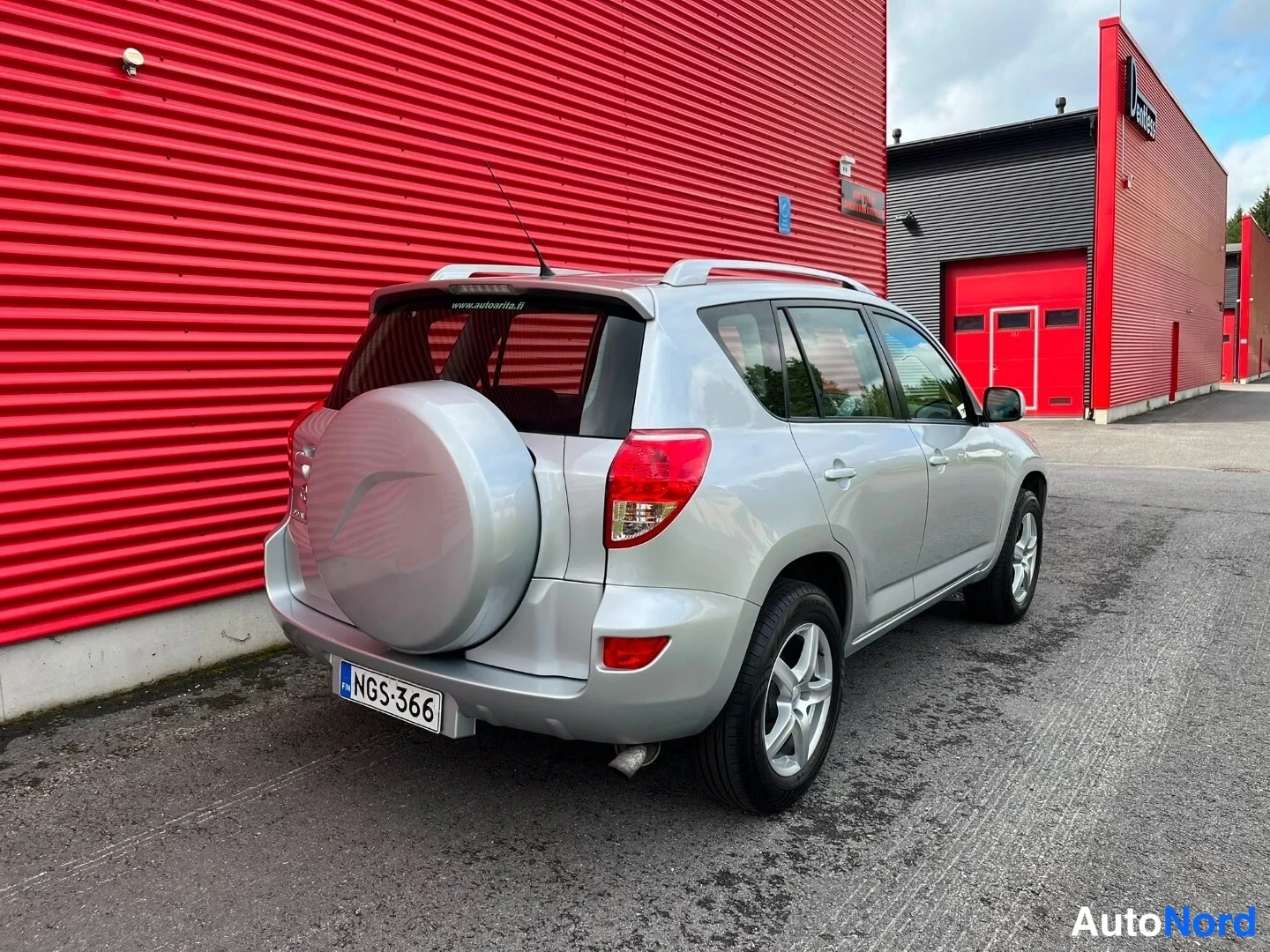 Takaviisto: Hopeanharmaan Toyota RAV4:n takaosa ja oikea kylki kolmasosakulmassa, varapyörä kannella, punaisen hallin edessä.