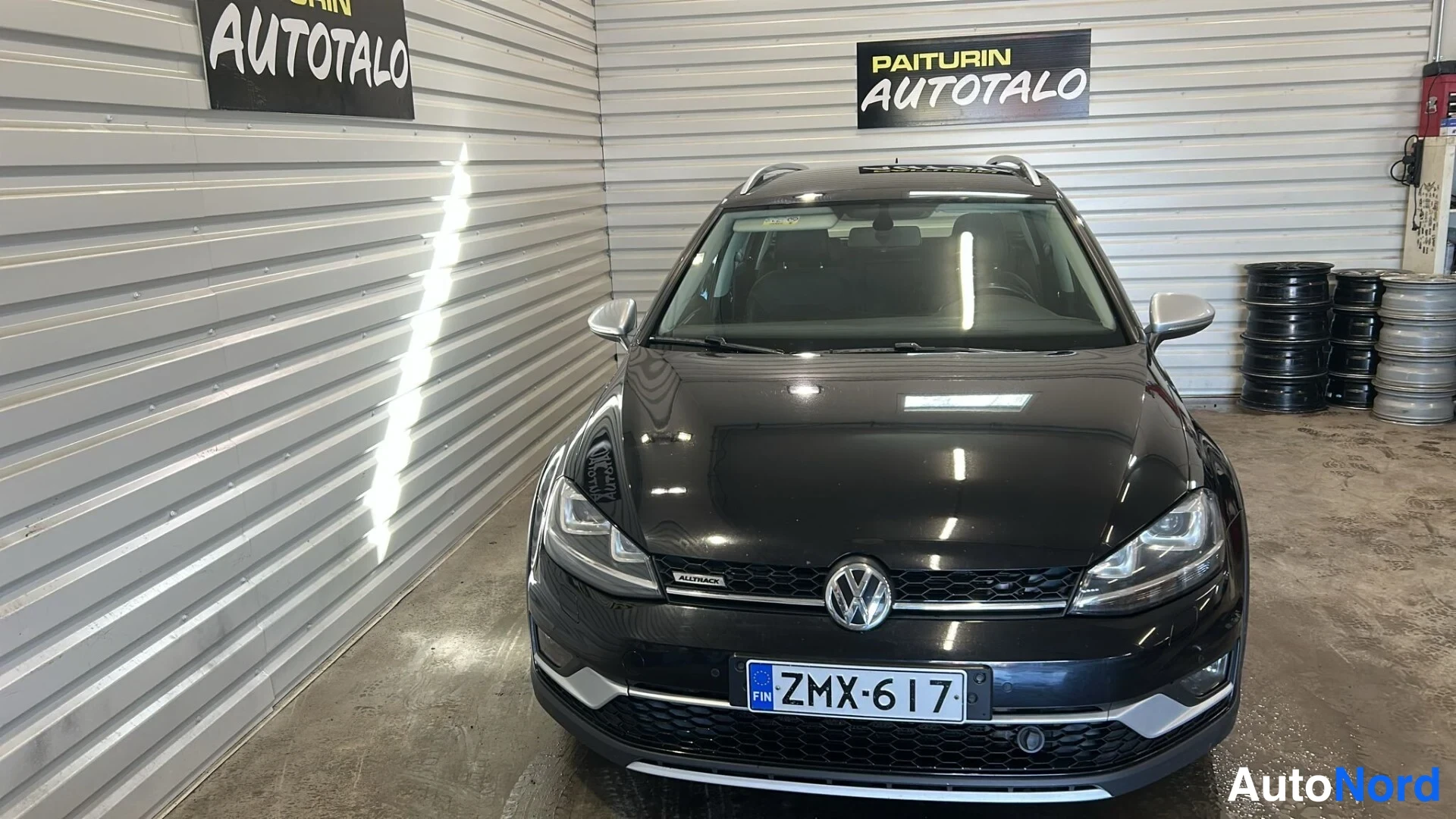 Edestä: Mustan auton etunäkymä sisätiloissa, VW-merkki ja rekisterikilpi näkyvät, renkaita kasassa taustalla.
