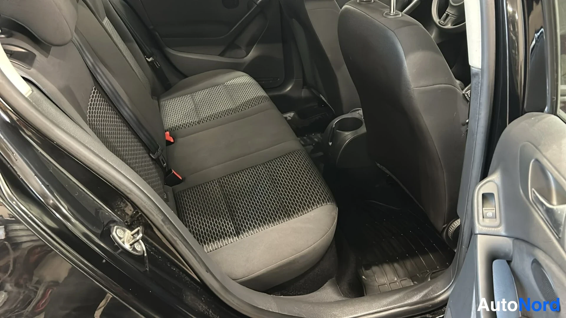Muu: Autojen takapenkki tummalla kankaanverhoilulla kuvattuna avoimesta takaovesta; keskikonsolissa kupinpitimet ja lattialla tumma matto.
