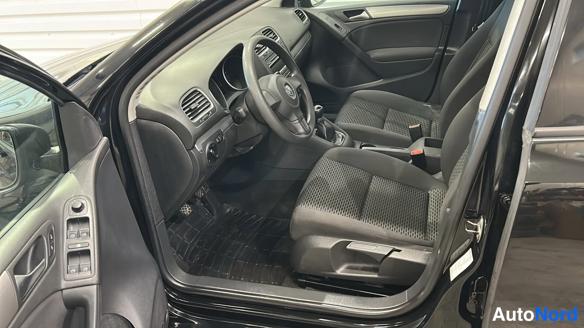 Edestä: Volkswagenin etuistuin- ja ohjaamotila kankaistuilla istuimilla ja mustilla mattoilla, kuljettajan ovi auki.