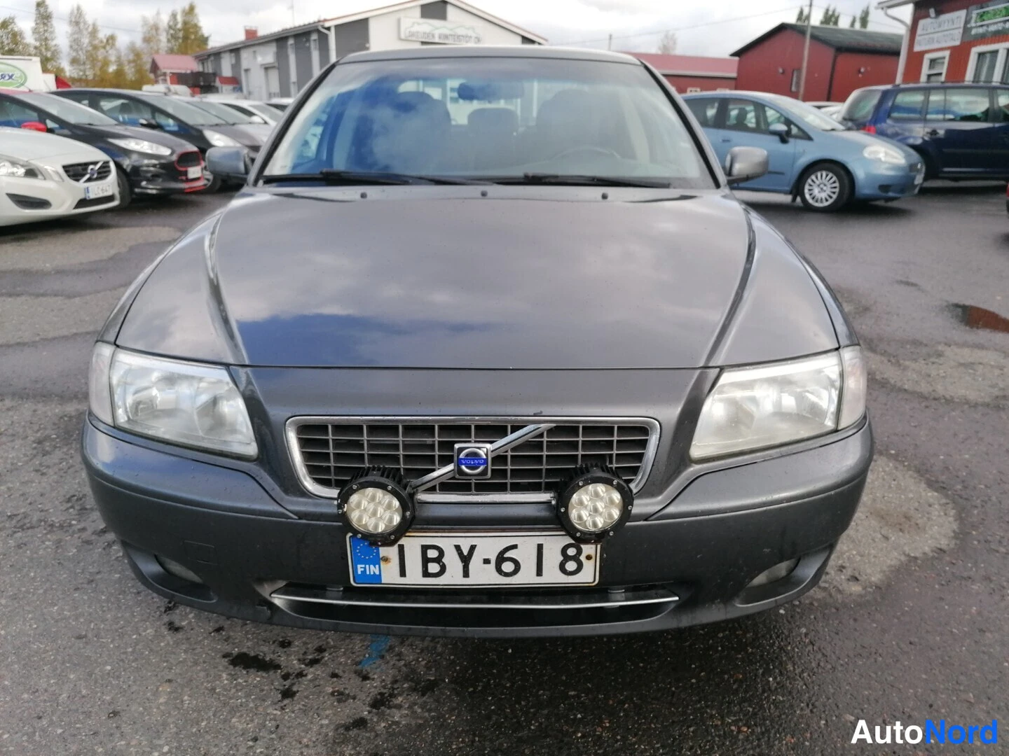 Edestä: Tummanharmaa sedan suora etunäkymä;Volvo-merkki keskeisesti säleikössä, kaksi pyöreää lisävaloa rekisterin yläpuolella, rekisteri.