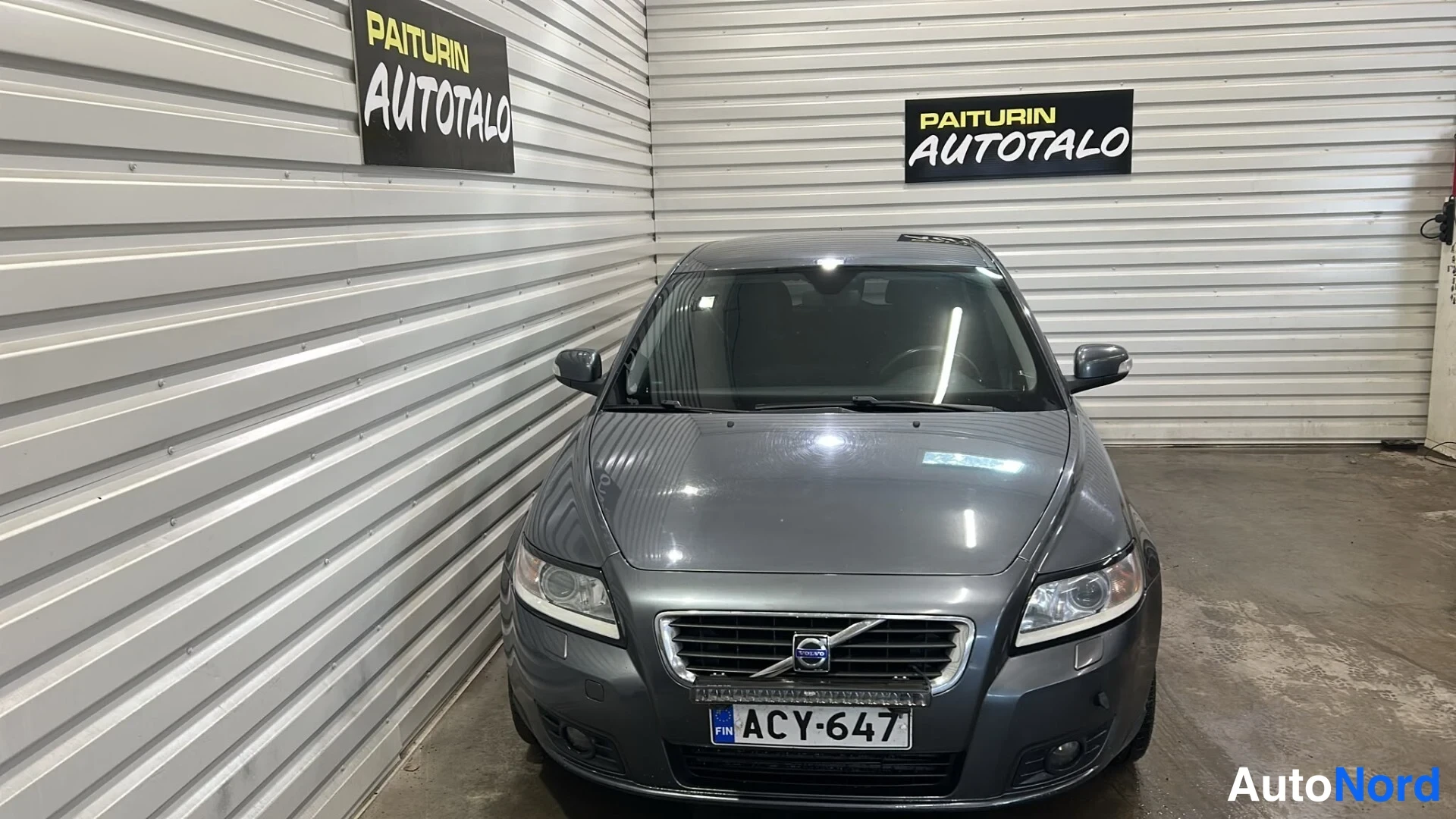 Edestä: Auton etupää suoraan edestä; tummanharmaa auto, Volvon etusäleikkö ja merkki sekä rekisterikilpi näkyvät.