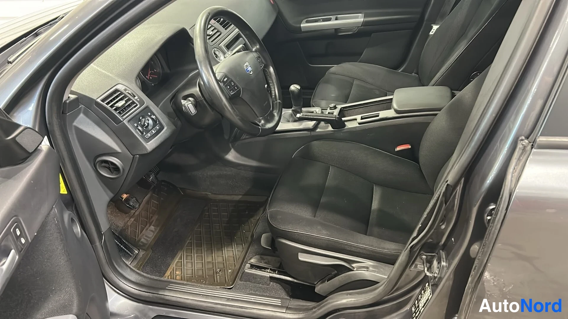 Muu: Avoimen kuljettajan oven näkymä Volvo V50:n etumatkustustilaan: musta ohjauspyörä, kangasistuimet, manuaalivaihteen vaihdekeppi ja kumimatot.