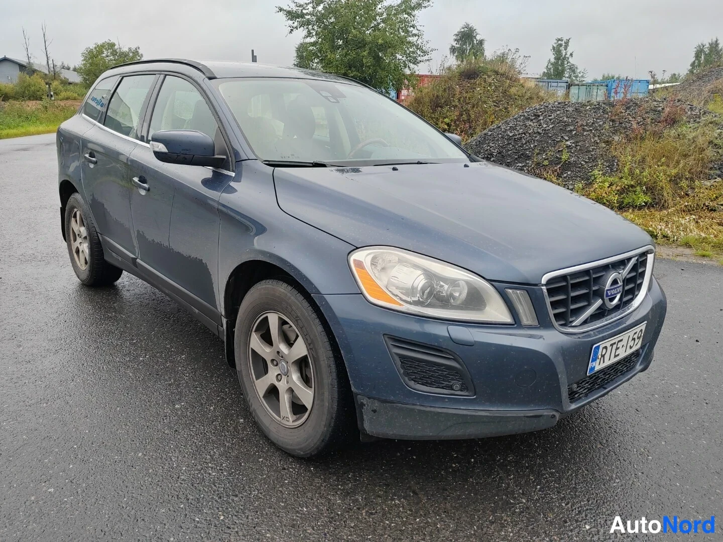 Etuviisto, nokka oikealle: Sinisenharmaan Volvo XC60:n etukolmaskuva; etupaneeli, ajovalot ja keula näkyvät märällä asfalttipihalla.