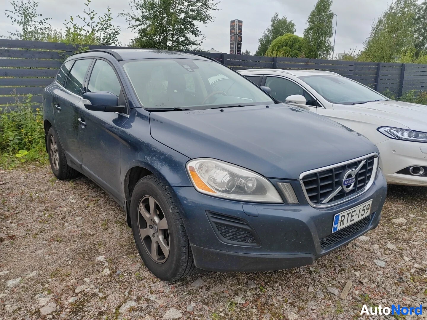 Etuviisto, nokka oikealle: Sininen henkilöauto ja valkoinen auto vieressä, edessä rekisterikilpi, sora- ja nurmipinta, takana puuaita.