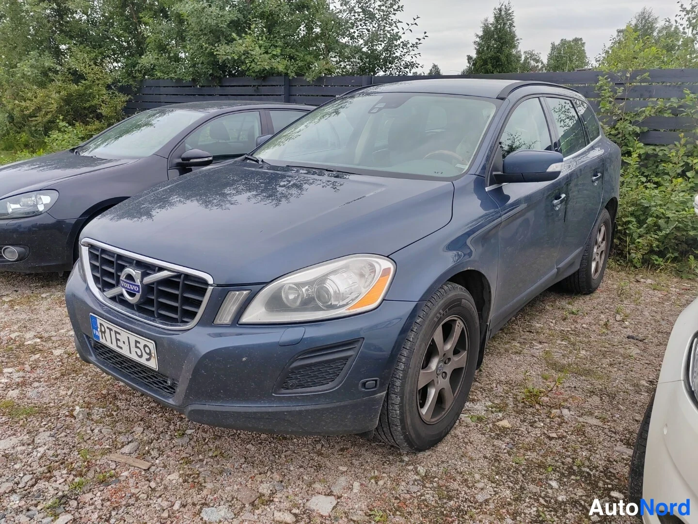 Etuviisto, nokka vasemmalle: Tummanharmaa tai sinertävä Volvo XC60 -maasturi kuvattu kolmeneljänneksestä edestä vasemmalle kääntyneenä sorapinteessä, rekisteritunnus.