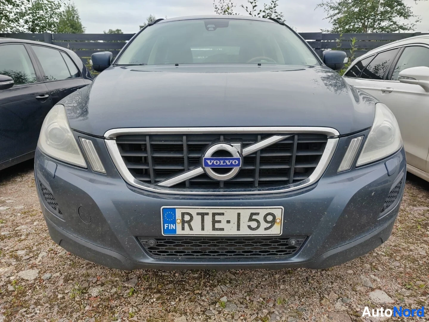 Edestä: Volvo XC60:n etupää suoraan edestä: VOLVO-merkki jäähdyttimensuojassa, FIN-tunnus Rekisterikilpi, tummanharmaa kori.