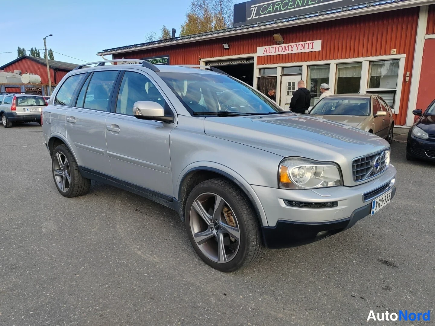 Etuviisto, nokka oikealle: Hopeanharmaa Volvo XC90 etuviistossa oikealta kuvattuna autotalon pihalla, keulakoriste ja etupyörät näkyvät.