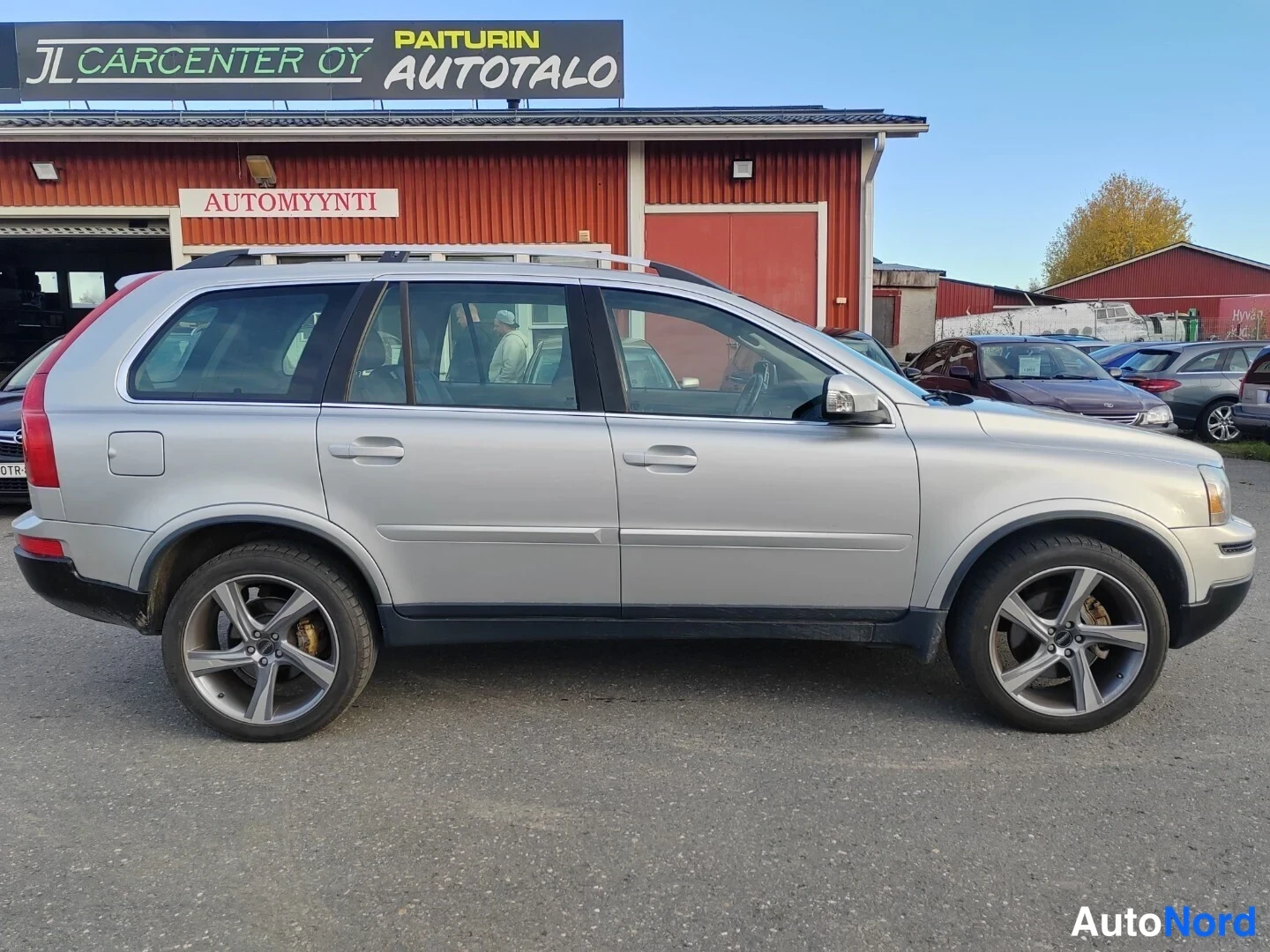 Sivulta: Hopeanharmaan Volvo XC90:n sivuprofiili, molemmat oviparit ja kattokaiteet näkyvät automyymälän edessä.