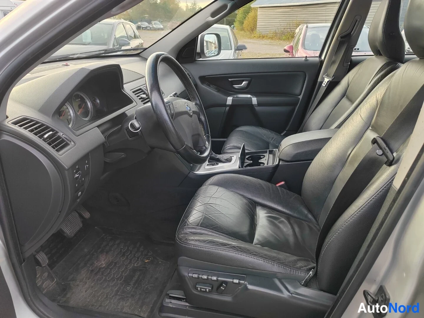Muu: Volvo XC90:n sisätila: musta nahkapenkit, ohjauspyörä ja mittaristo näkyvät kuljettajan ovesta kuvattuna.
