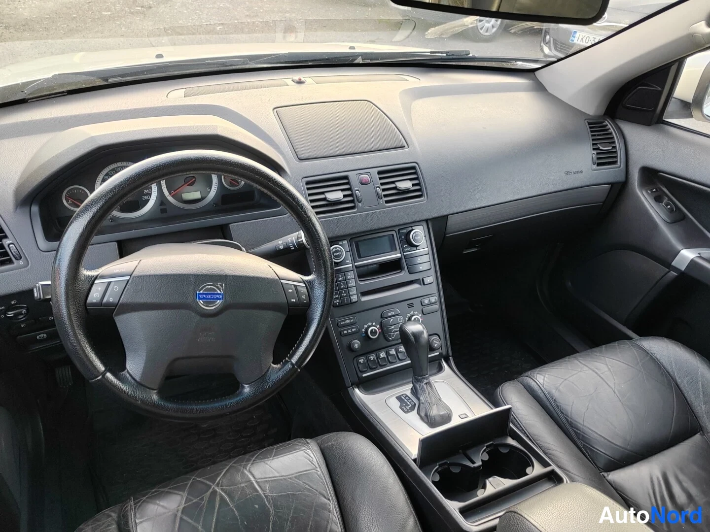 Muu: Volvo XC90:n ohjaamo vasemmalta: Volvon logolla varustettu ohjauspyörä, mittaristo, keskikonsoli ja automaattivaihteen valitsin, mustat nahkaistuimet.