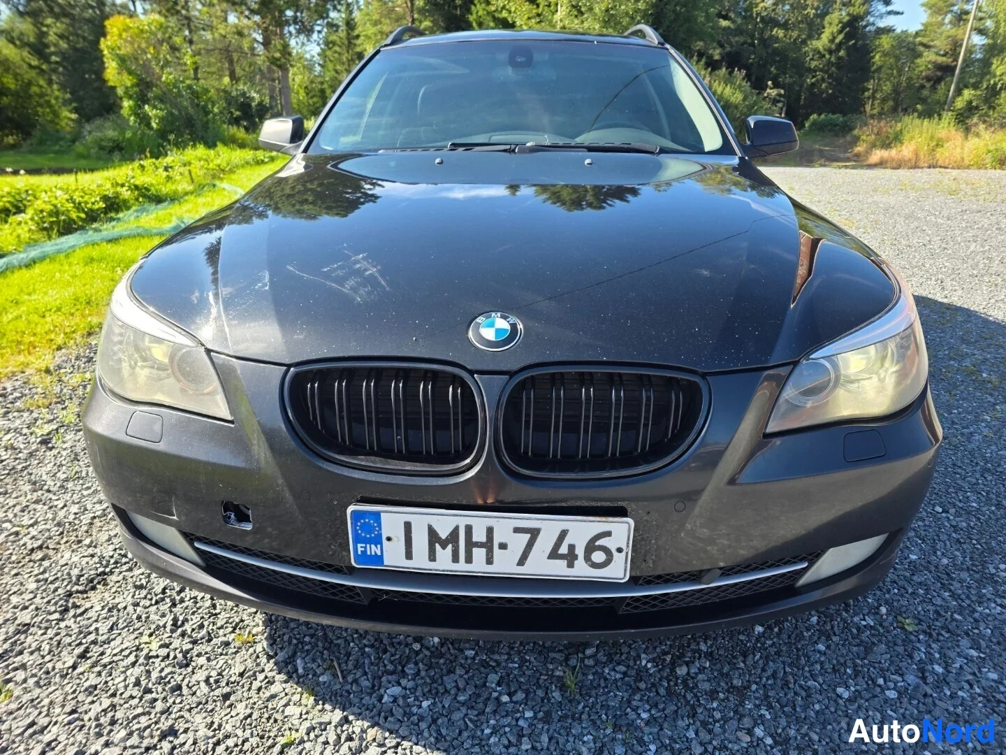 Edestä: Mustan auton etupää suoraan edestä, BMW-merkki jäähdyttäjäsuodattimessa, rekisteritunnus, maalipinnassa naarmuja konepellillä.