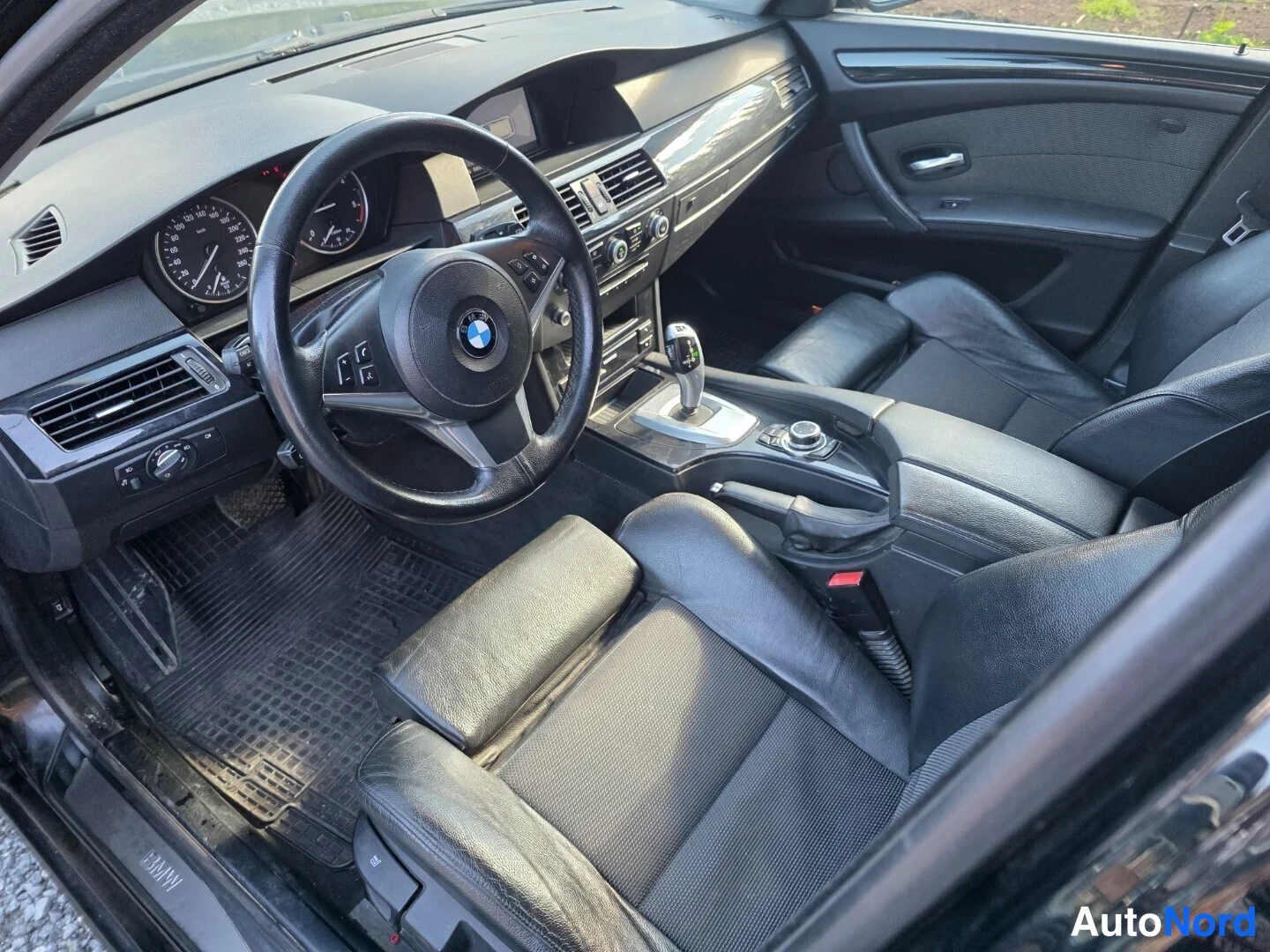 Edestä: Auton sisätilat ohjaamosta kuvattuna: BMW-ohjauspyörä, mittaristo, manuaali-ilmastointilaitteet ja automaattivaihteen valitsin, mustat istuimet ja ovipaneelit.