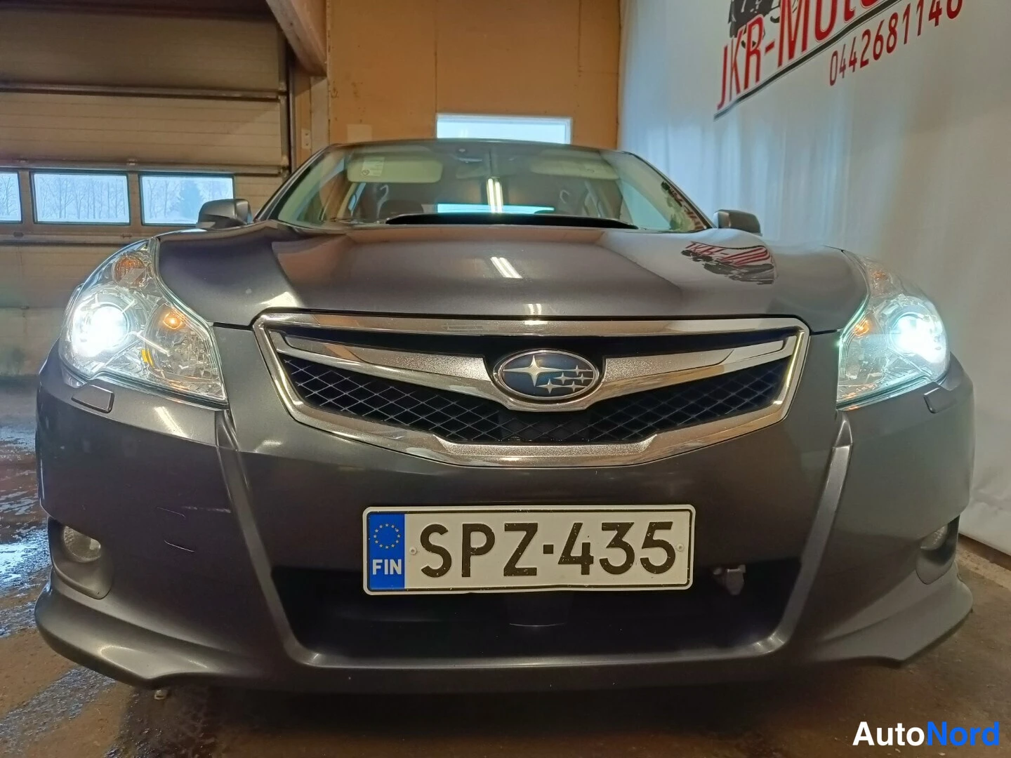Edestä: Tummanharmaan sedanin keula suoraan edestä, Subaru-merkki etusäleikössä ja rekisterikilpi.