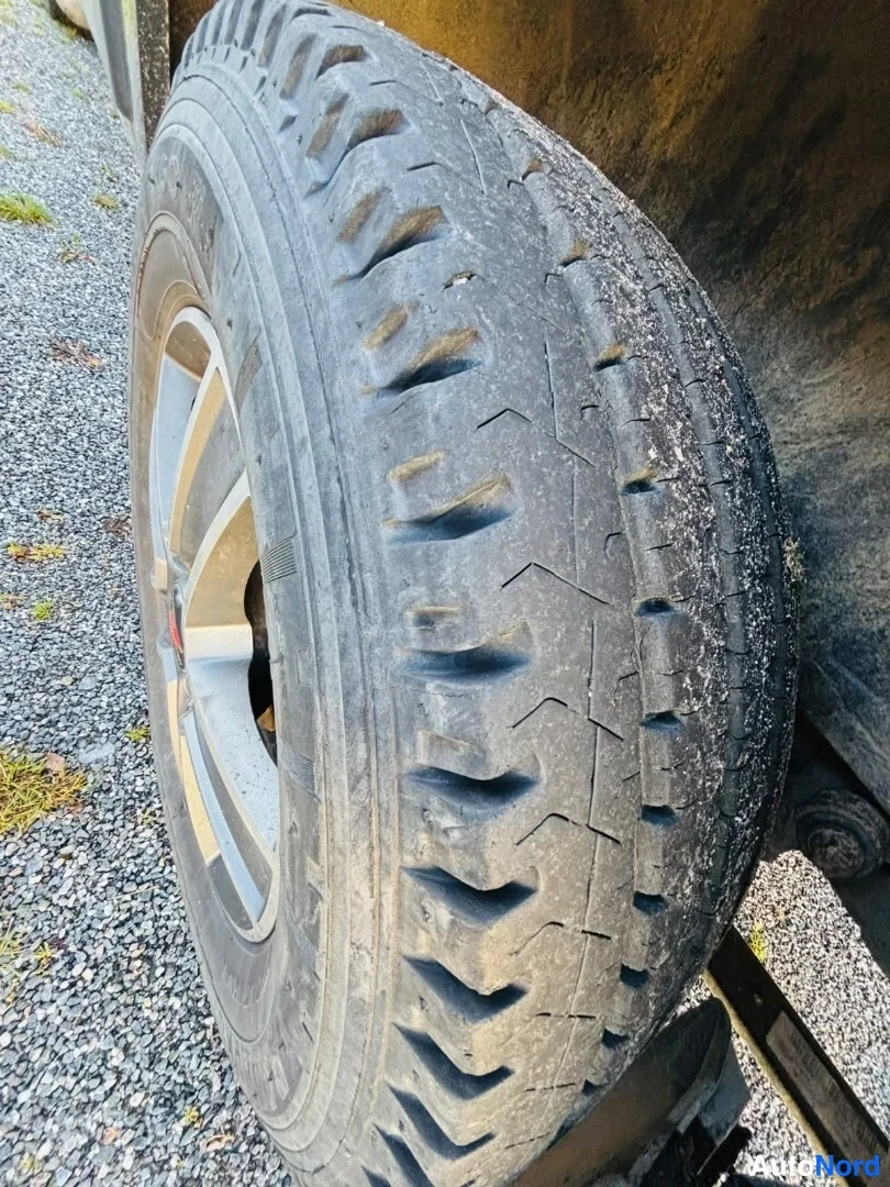 Yksityiskohta: Lähikuva auton takarenkaasta ja osasta alumiinivannetta sorapäällysteisen alustan päällä; kuluneen näköinen nasta-urituinen kesärenkaan kulutuspinta.