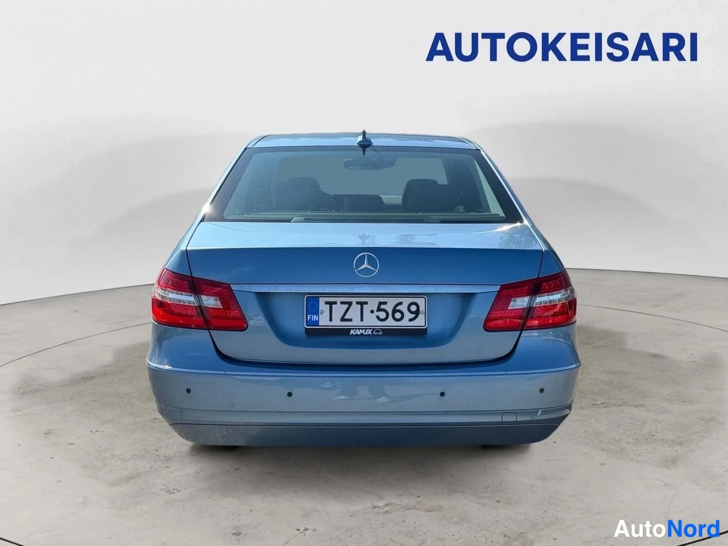 Takaa: Sininen Mercedes-Benz E-sarjan sedan suoraan takaa kuvattuna, perävalot, kannenlatti ja rekisterikilpi näkyvissä studiossa.