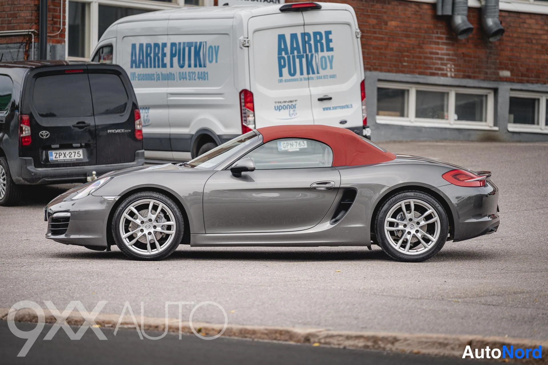 Sivulta: Porsche Boxster avomalli punaisella katolla kuvattu sivusuunnasta; auton kylki vasemmalla puolella, tausta autoja lähikuvissa.