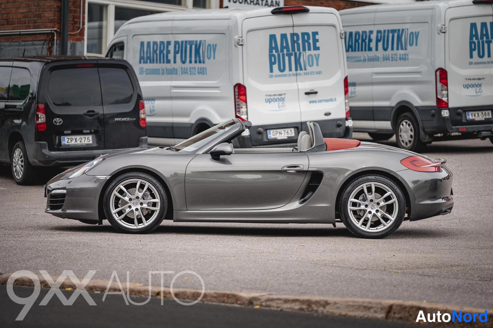 Etuviisto, nokka vasemmalle: Porsche Boxster-avoneva front three-quarter -kuva vasemmasta etukulmasta, katto auki, kori harmaa, punainen sisätilakansi näkyvissä.
