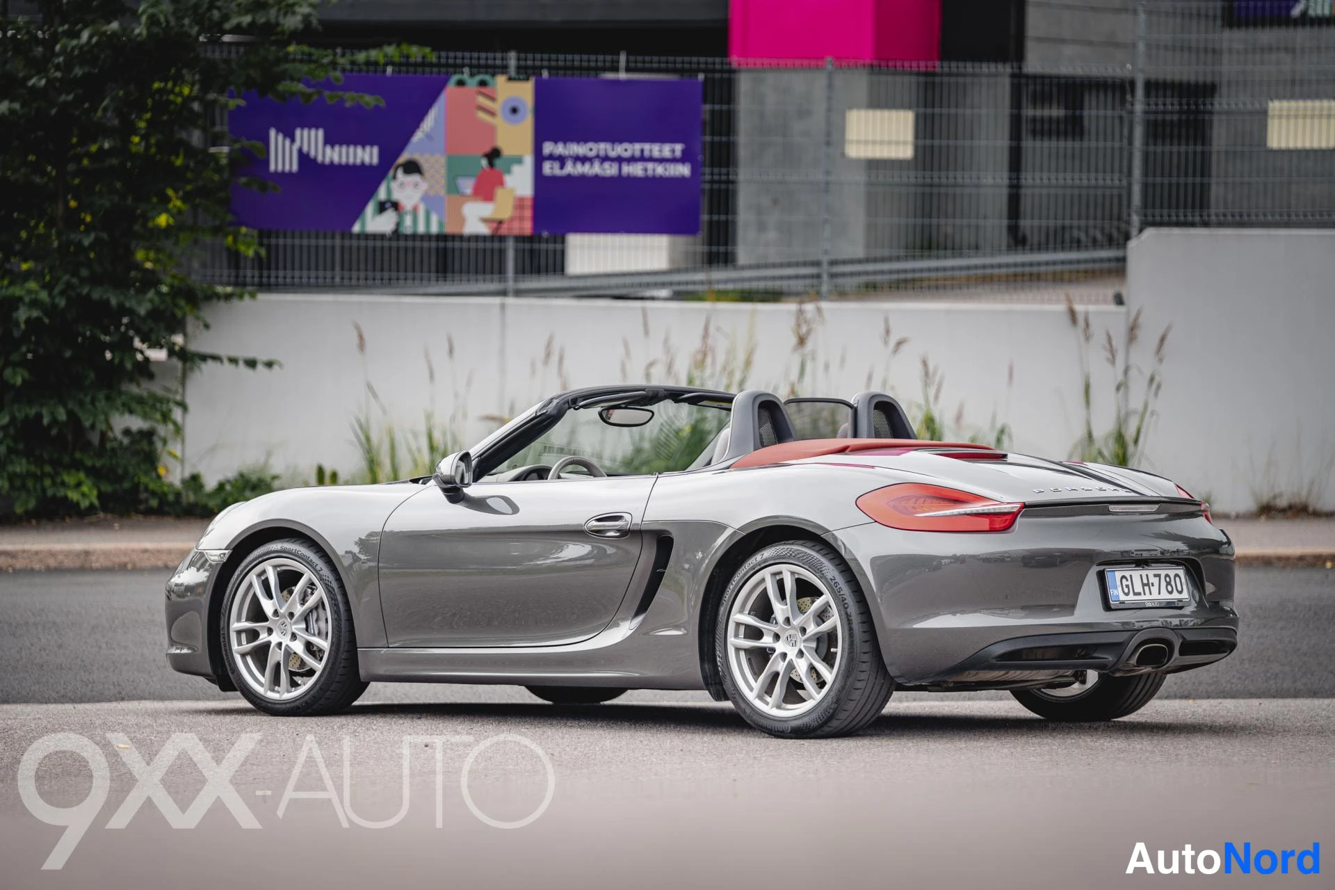 Takaviisto: Porsche Boxster-avoneva rear three-quarter -kuva vasemmasta takakulmasta, katto auki, punainen kansi ja rekisteri näkyvissä.