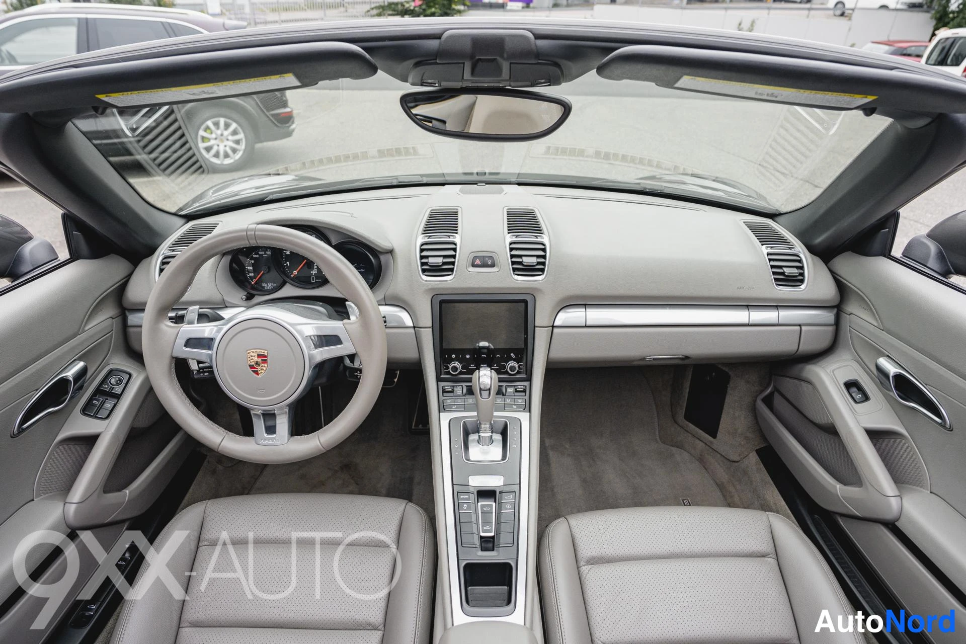 Takaviisto: Porsche Boxsterin ohjaamo ylhäältä: beige nahkapaneelit, Porsche-ohjauspyörä ja keskikonsolin näyttö.