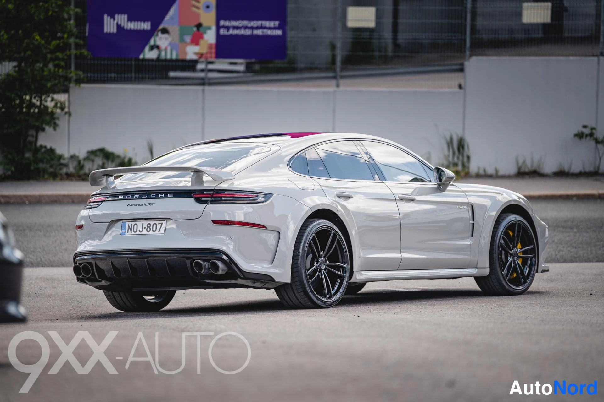 Takaviisto: Hopeanharmaa Porsche Panamera takaviistosta, takasiipi ylhäällä, mustat vanteet ja keltainen jarrusatula näkyvissä, rekisteritunnus.
