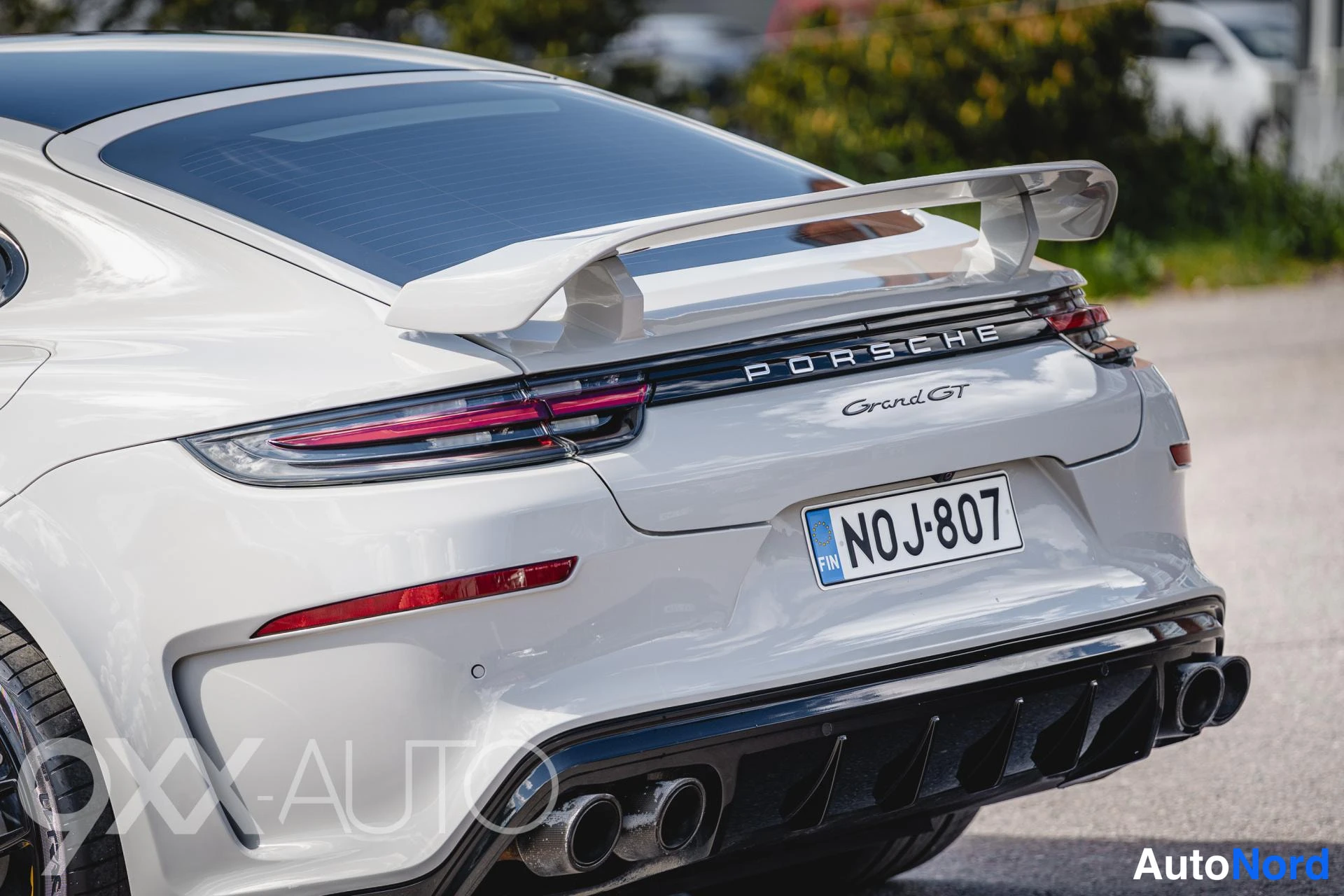 Takaa: Lähikuva vaalean auton takaosasta, PORSCHE Grand GT -teksti takaosassa, takasiipi ja neljä pakoputkea, rekisteritunnus.
