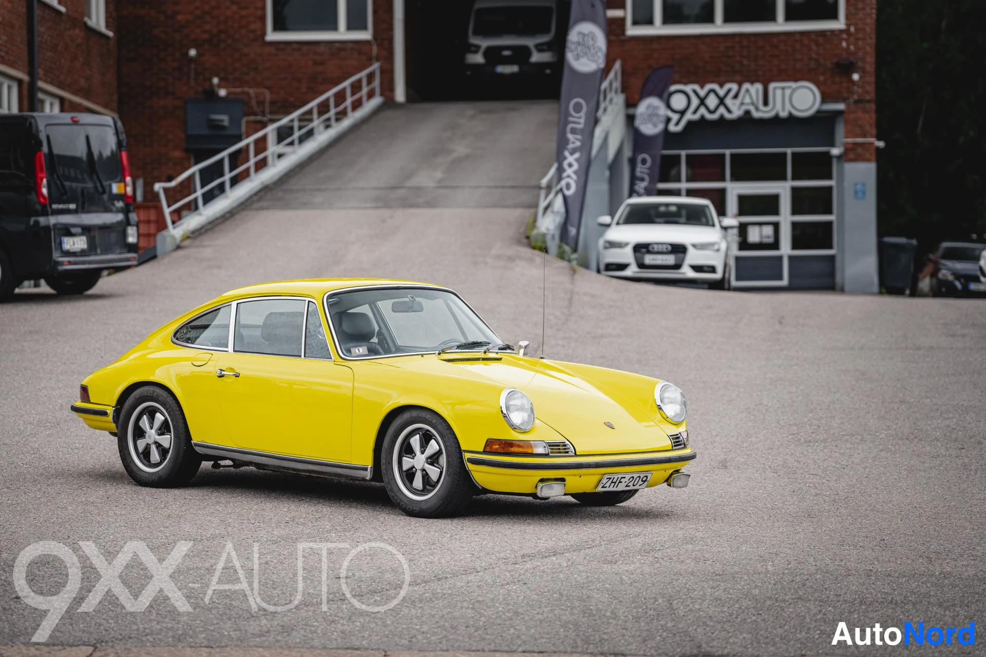 Etuviisto, nokka oikealle: Keltainen klassinen Porsche-urheiluauto kolmivartaloisenä etuviistokuvassa, nokka oikealla, parkkipaikalla autotallin edustalla.