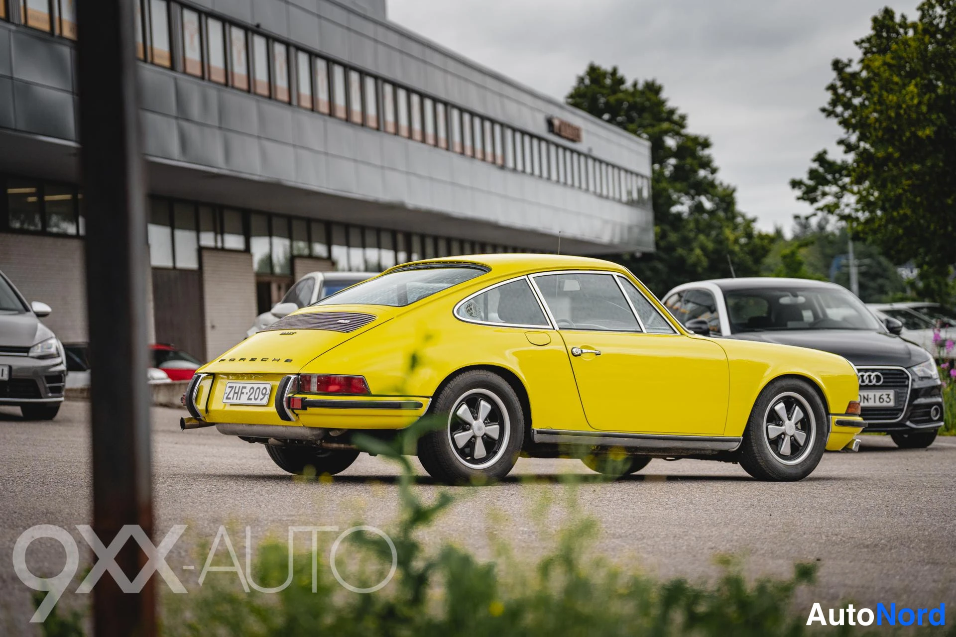 Takaviisto: Keltainen Porsche 911 -coupe takaviistosta vasemmalta; takaosa, rekisterikilpi, PORSCHETEKSTI ja peltivanteet näkyvät, taustalla rakennus ja muita autoja.