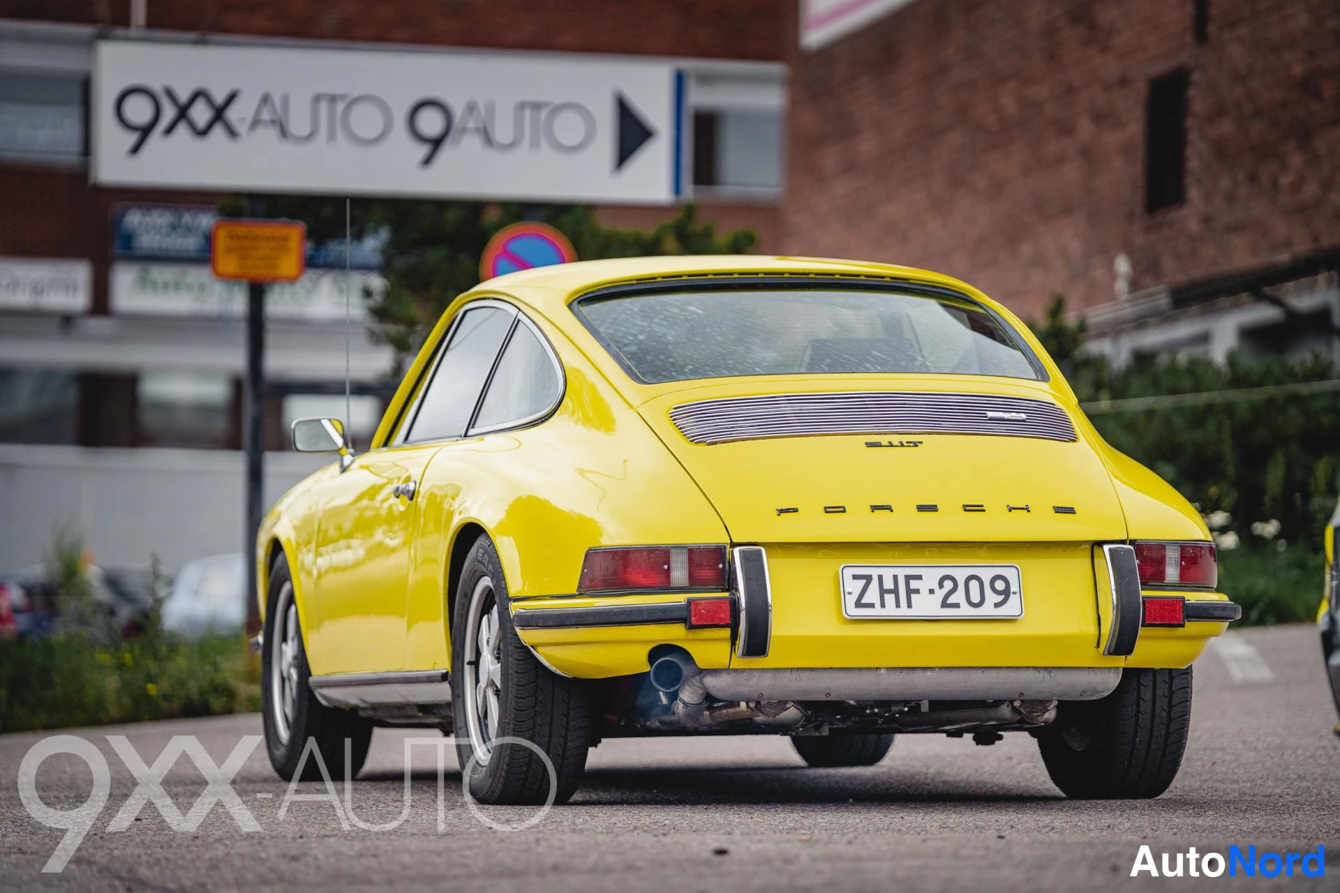 Takaviisto: Keltainen Porsche 911 -coupe takaa vasemmalta kuvattuna; takalasi, ritilä, PORSCHE-teksti, -kilpi ja kaksoisputki näkyvät. Taustalla 9XX-AUTO-kyltti.