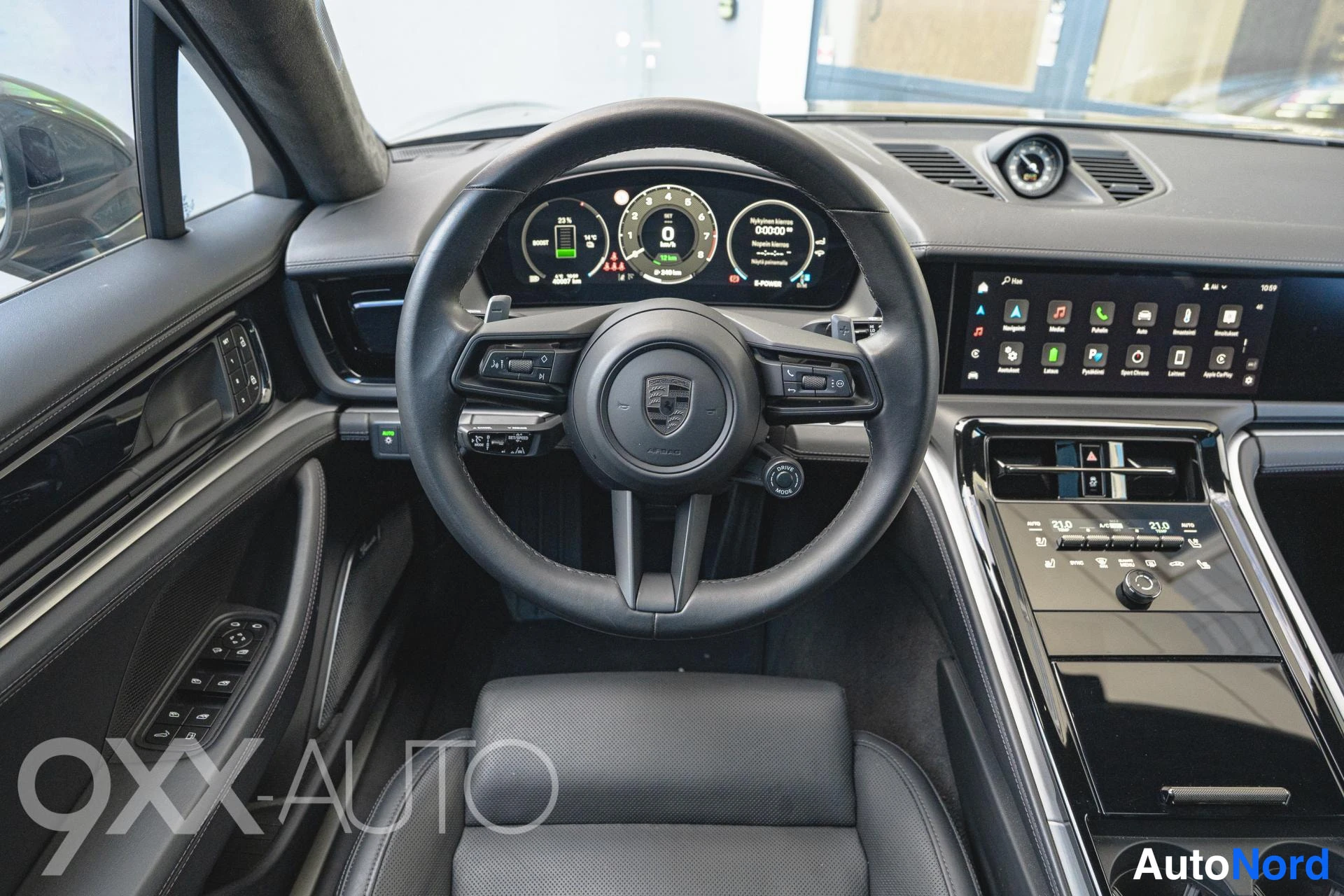 Etuviisto, nokka oikealle: Autojen sisätilat: Porsche-ohjauspyörä, digitaalinen mittaristo (lukema 00000 km) ja keskinäyttö sovelluskuvakkeineen, musta sisusta ja penkit.