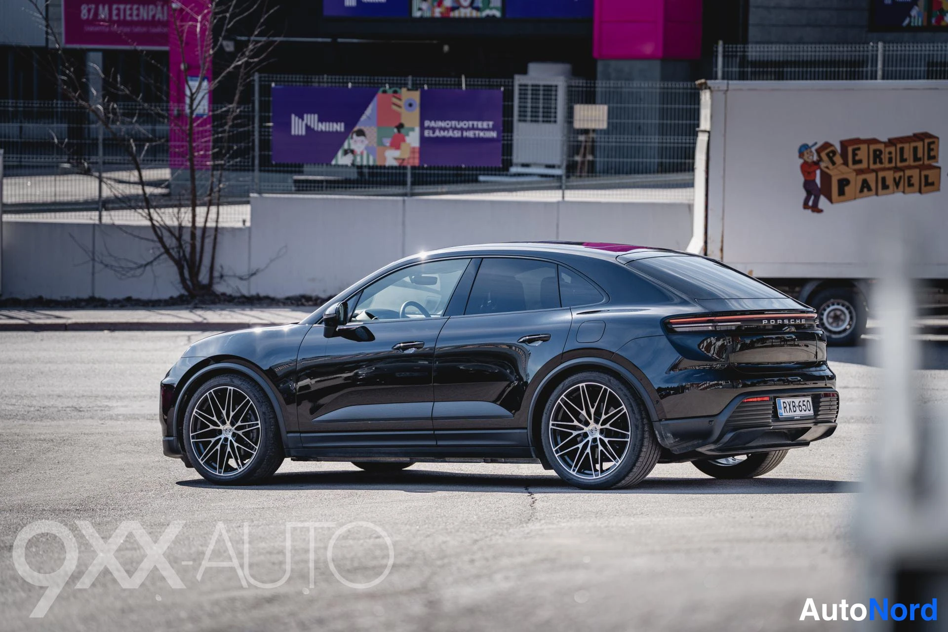 Takaviisto: Musta Porsche Macan -tyyppinen SUV takaviistosta, rekisterikilpi, kaupunkiympäristössä.