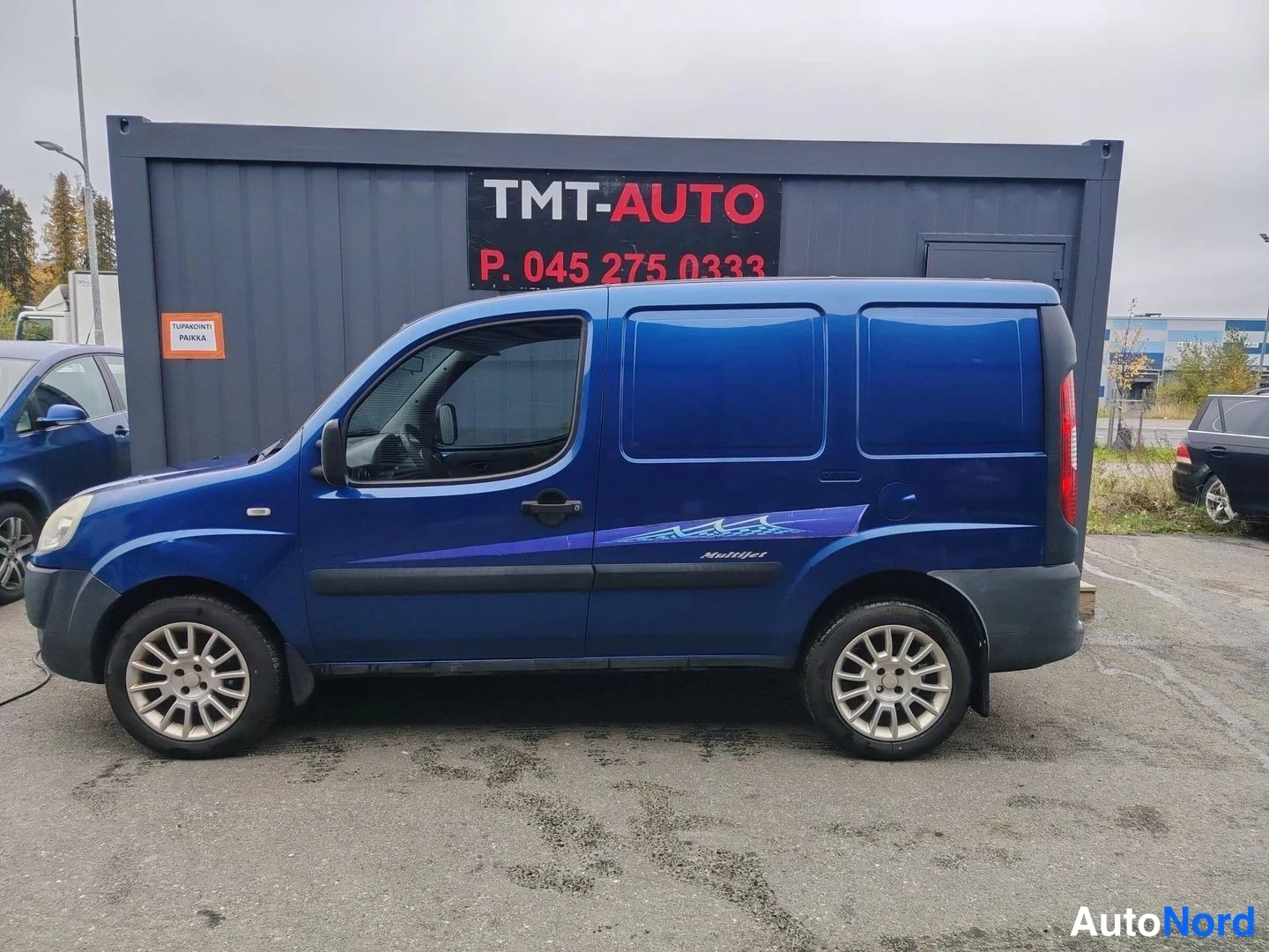 Sivulta: Sininen Fiat Doblo sivulta, rekisterikilpi ei näy, TMT-Auto-kontti ja autopaikka-taulu taustalla.