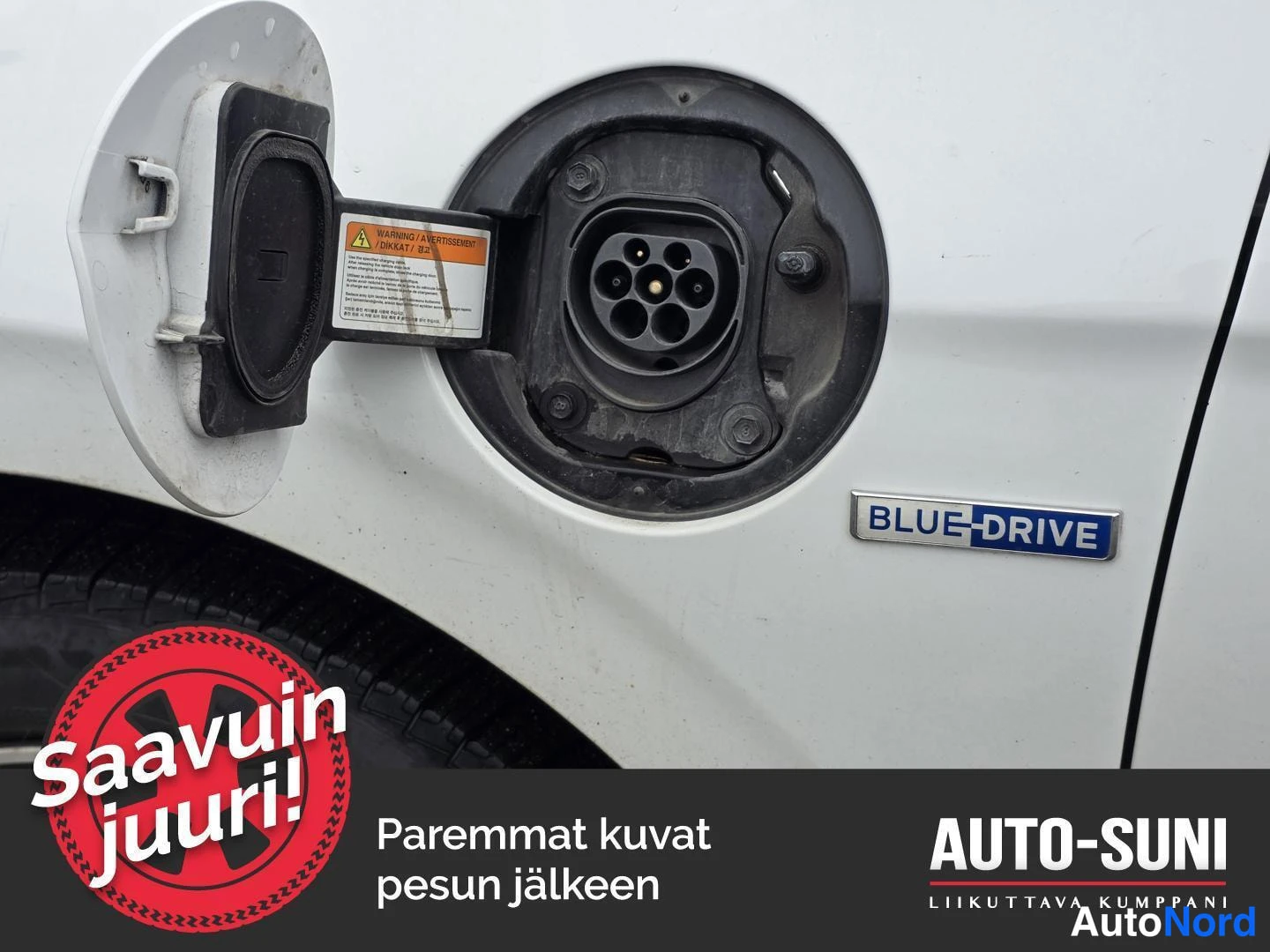 Yksityiskohta: Auton latausportti auki. Latausliitin ja sininen BLUE-DRIVE -logo ruudussa. Osa renkaasta näkyvissä alareunassa.