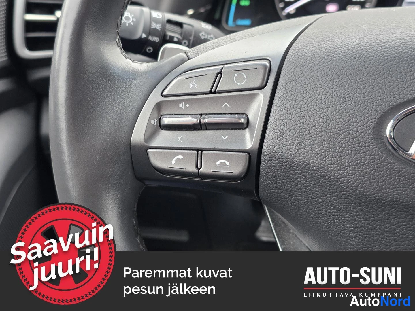 Sivulta: Sisäkuva ohjauspyörän vasemman sivun painikkeista: puhekomennon ja puhelun painikkeet sekä äänenvoimakkuussäädin; taustalla osittain näkyvä mittaristo ja AUTO-m