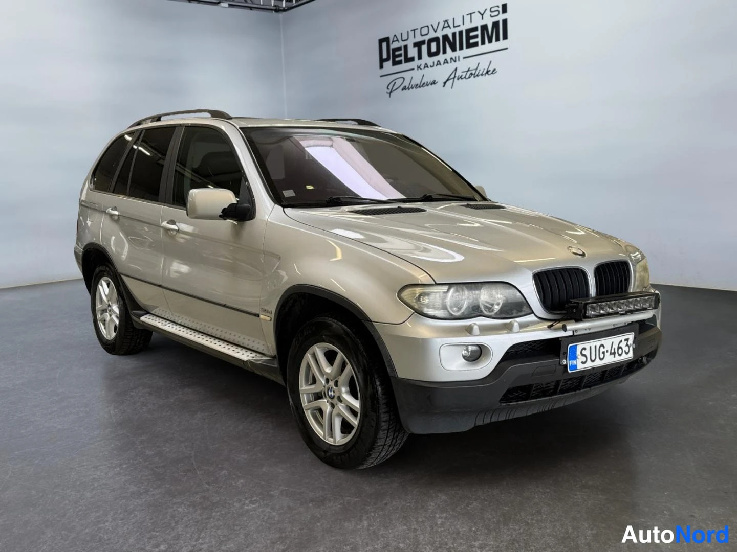Etuviisto, nokka oikealle: Hopea BMW X5 -maastauto etukulmasta oikealta, rekisteritunnus, auton keulassa lisävalo, studiossa.
