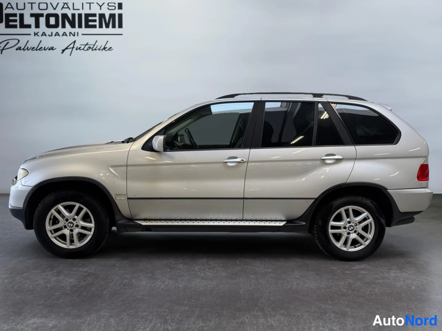 Sivulta: Sivukuva hopeanvärisestä BMW X5 -maasturista sisätiloissa; molemmat ovet, kattokaiteet ja sivutekot näkyvät.