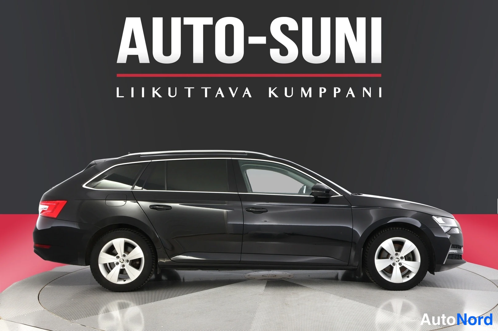 Sivulta: Side profile view of a dark grey Skoda Superb station wagon showing the driver's side. Features visible are tinted windows, body-colored door handles, and silve