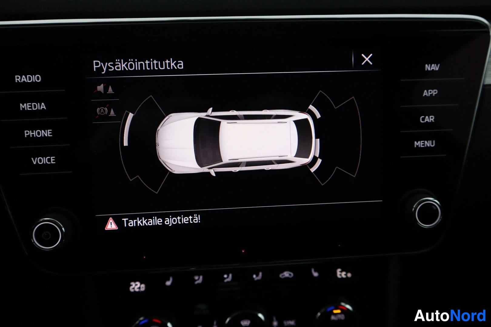 Yksityiskohta: Infotainment-järjestelmän näyttö, jossa esitetään 'Pysäköintitutka' ja auton päällinen karttapohjaisena mallina. Varoitusviesti 'Tarkkaile ajotietä!' ja huomio