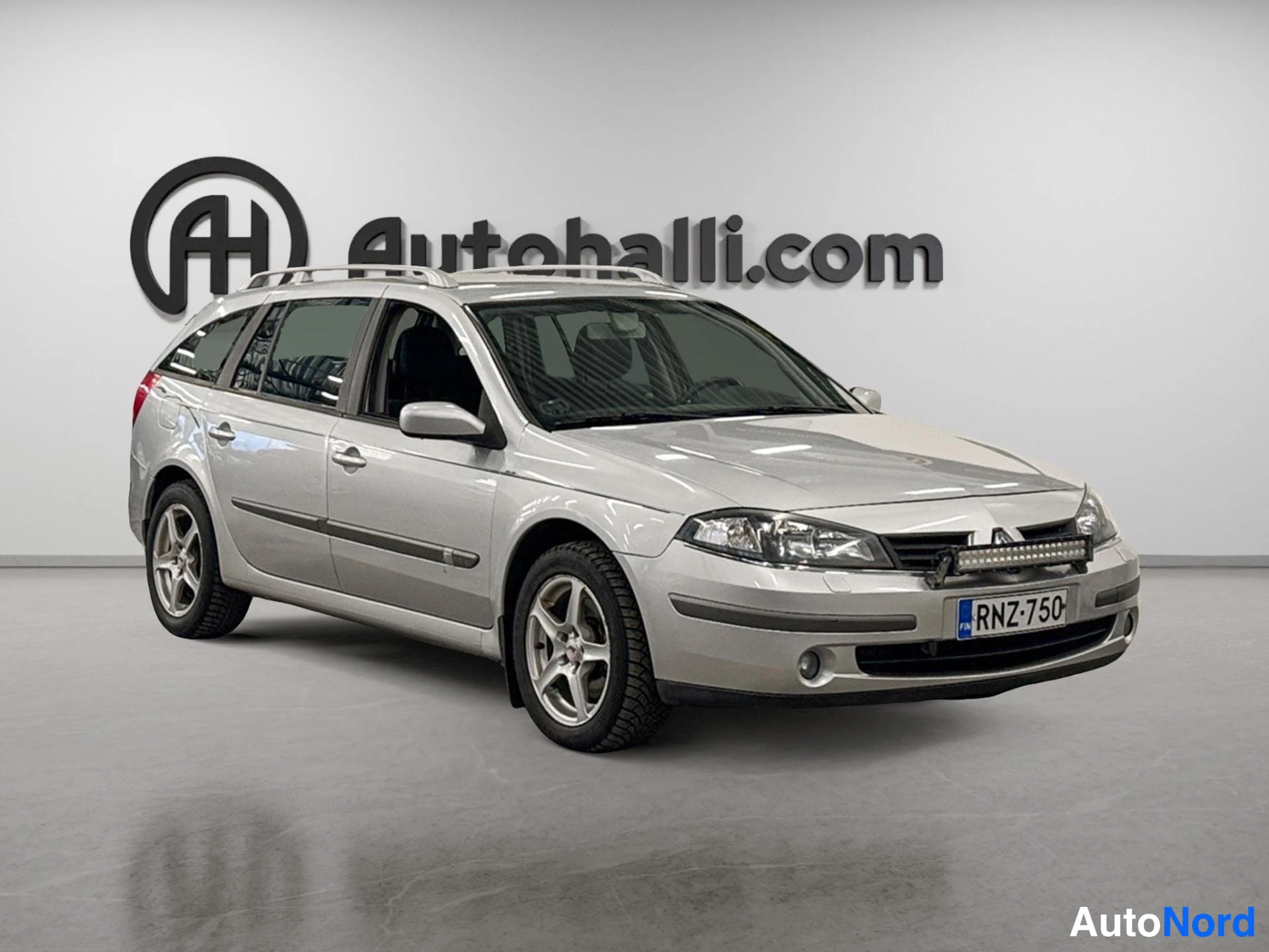 Etuviisto, nokka oikealle: Hopeanharmaa Renault Laguna -farmari etuosa oikealta vasemmalle kuvattuna; konepellillä ja etupuskurissa lisävalo, rekisteri.