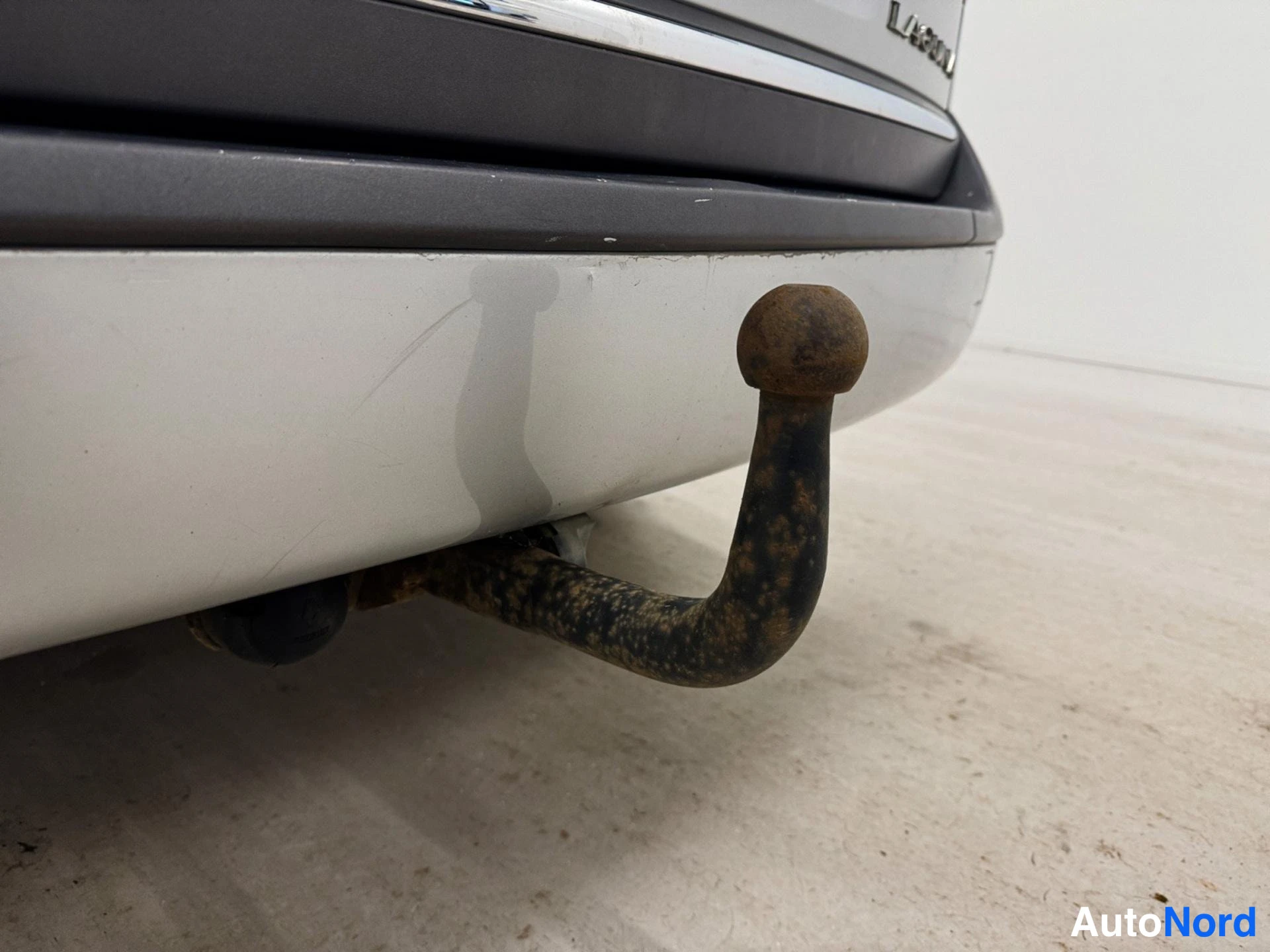 Yksityiskohta: Lähikuva autojen vetokoukusta. Mustan muovipuskurin alapuolella on ruosteinen metallinen vetokoukkupuu valkoisessa puskurissa.