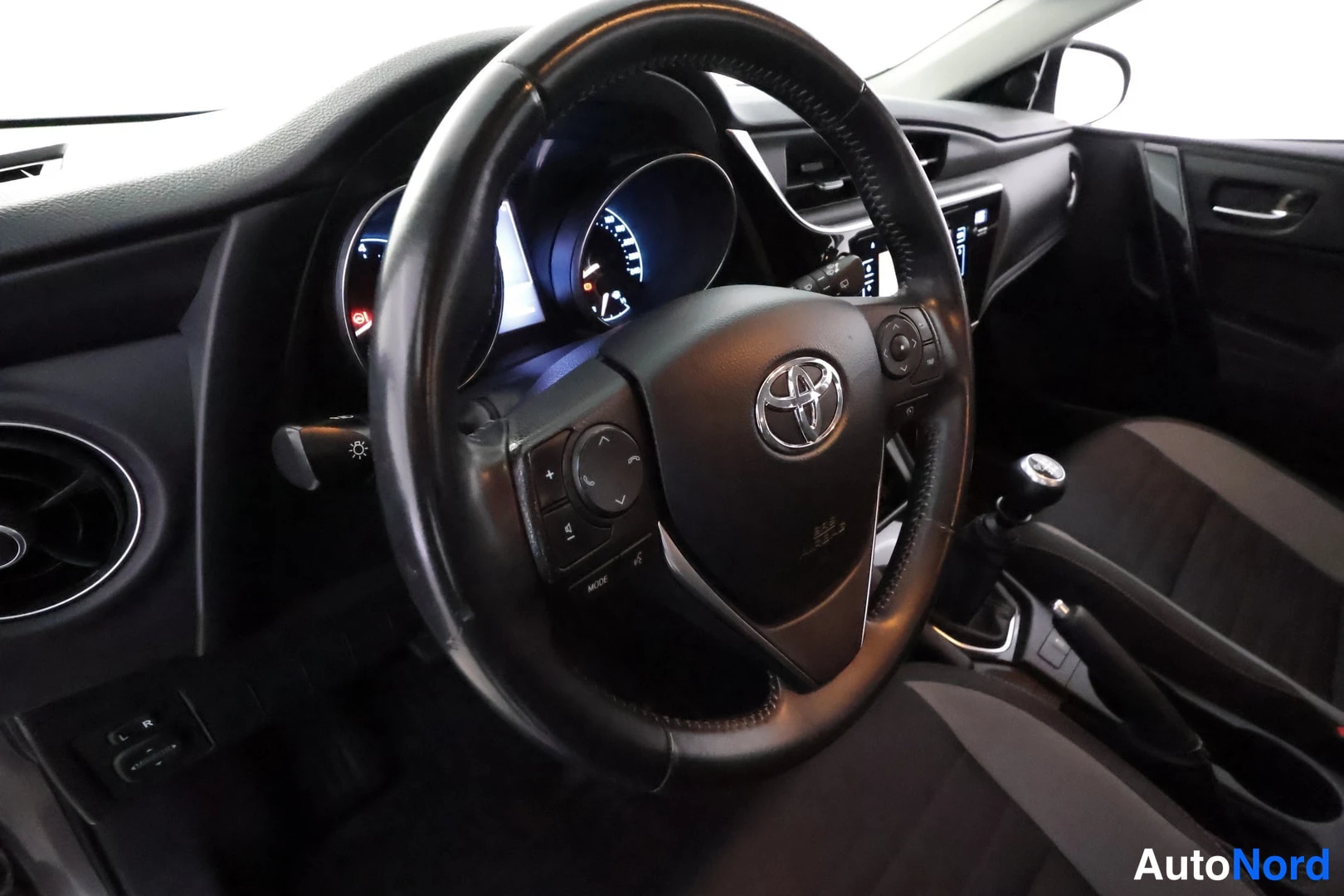 Muu: Sisäkuva: Toyota-merkkinen ohjauspyörä, kojelaudan mittarit ja manuaalivaihteistoon viittaava vaihdekeksi.