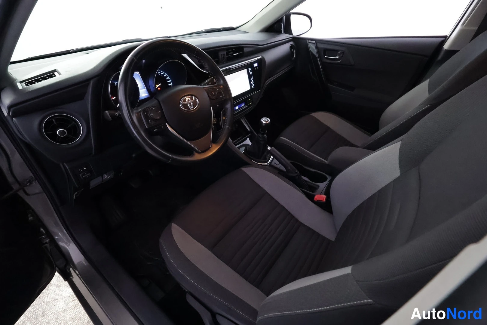 Muu: Toyota Aurisin ohjaamo vasemmalta ovilta kuvattuna: ohjauspyörä, mittaristo ja kosketusnäyttö näkyvät, vaihdossa on manuaalinen vaihteenkehys, etupenkit ovat ha