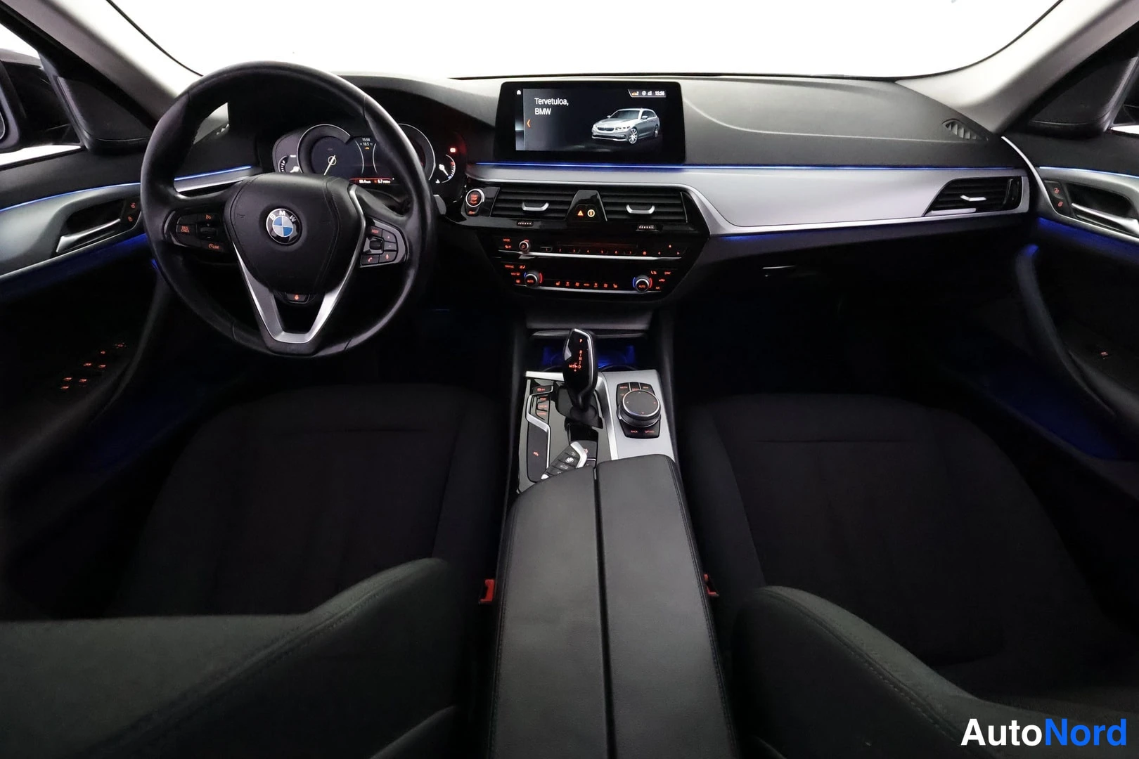 Sivulta: Kojetausta ja ohjaamon yleiskuva: BMW-tunnuksellinen ohjauspyörä, mittaristo, keskikonsoli ja infotainment-näyttö, jossa teksti 'Tervetuloa, BMW'.