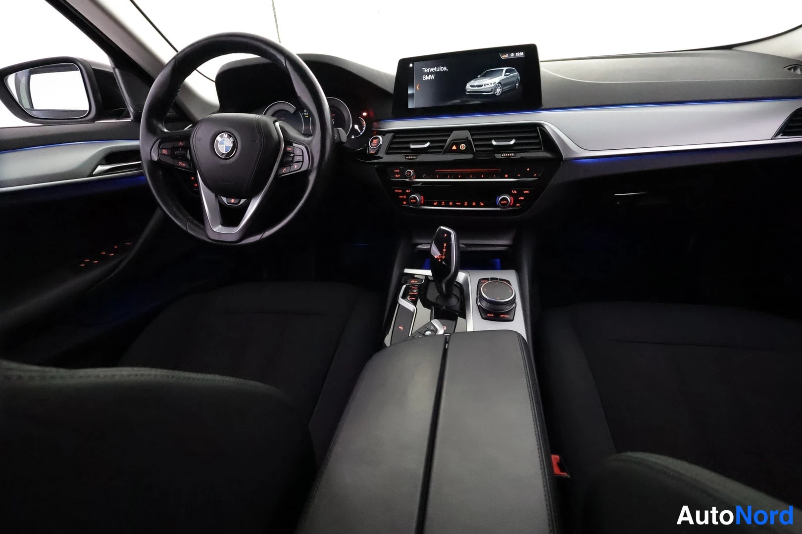 Sivulta: Ohjaamo kuvattuna takaistuimelta päin: BMW-ohjauspyörä, mittaristo, keskusnäyttö jossa teksti 'Tervetuloa, BMW', automaattivaihteen valitsin, iDrive-hallintapyö