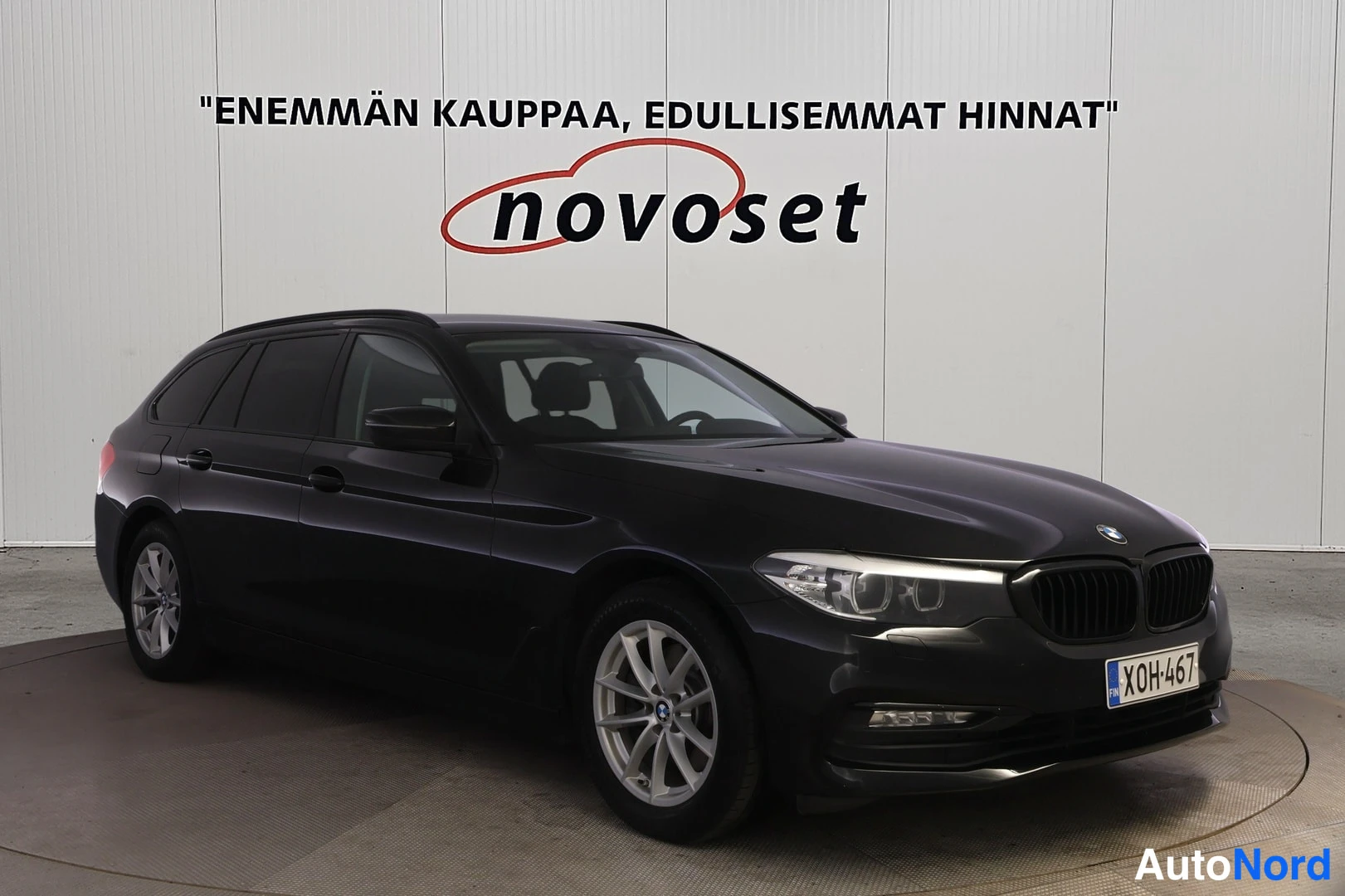 Etuviisto, nokka vasemmalle: Musta BMW 5 -sarjan farmari näkyy etukulmasta. Auto seisoo valkoisessa huoneessa ja taustalla on novoset-logo ja teksti ENEMMÄN KAUPPAA, EDULLISEMMAT H