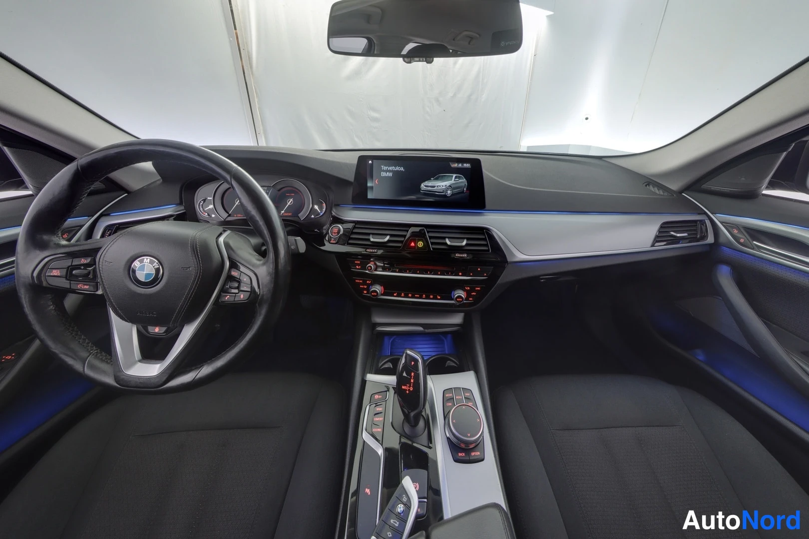 Ohjaamo: Ohjaamon näkymä: ohjauspyörä jossa BMW-logo, mittaritila ja keskusnäytössä sanoma Tervetuloa, BMW. Sininen sisävalaistus kirkastaa ohjaamon.
