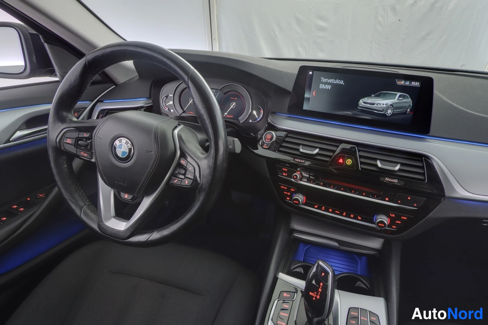 Ohjaamo: Ohjaamo lähikuva; musta nahkaohjauspyörä, mittaritaulu, navigaationäyttö jossa lukee Tervetuloa, BMW sekä automaattivaihteisto ja sininen sisävalo.