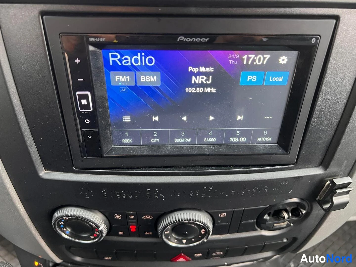 Yksityiskohta: Kojelaudassa Pioneer-kiintolevyradio (DMH-A240D), näytöllä Radio FM1, asema NRJ 102,80 MHz ja kello 17:07; alapuolella lämmityksen säätönupit ja painikkeet.